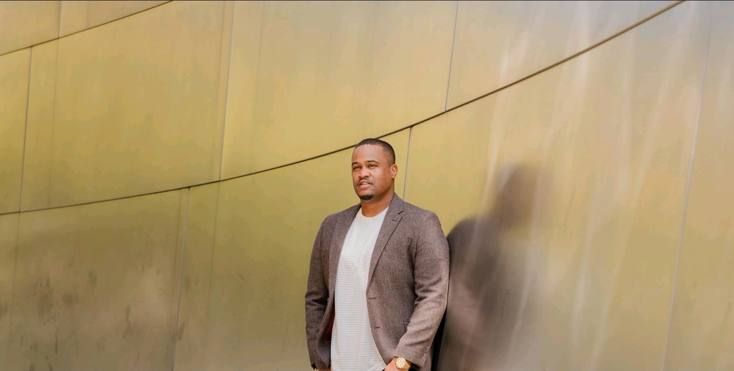 E'Ian West standing against a sculptural gold wall portrait backdrop E'Ian West standing against a sculptural gold wall portrait backdrop
